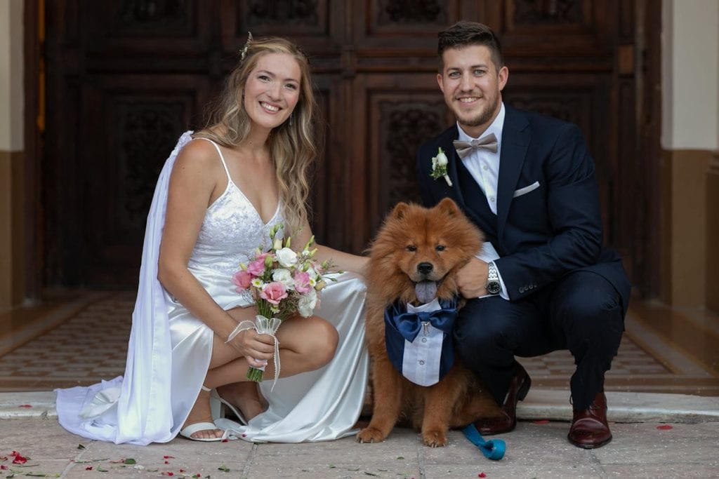 A bride and groom crouch and smile, posing with their brown, fluffy dog dressed in a blue tuxedo. The bride holds a bouquet and wears a white dress; the groom is in a dark suit with a bowtie. They are outdoors before a wooden door.