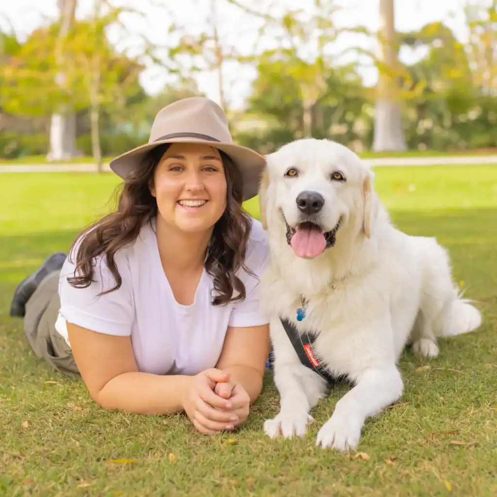 A woman in a white shirt and beige hat lies on the grass next to a large, happy white dog. Both are facing the camera, smiling, with trees and greenery in the background.