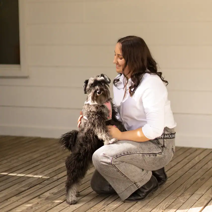 A woman kneels on a wooden deck, smiling as she hugs a small, fluffy black and gray dog. The background features a white wall and window, creating a warm and friendly atmosphere.