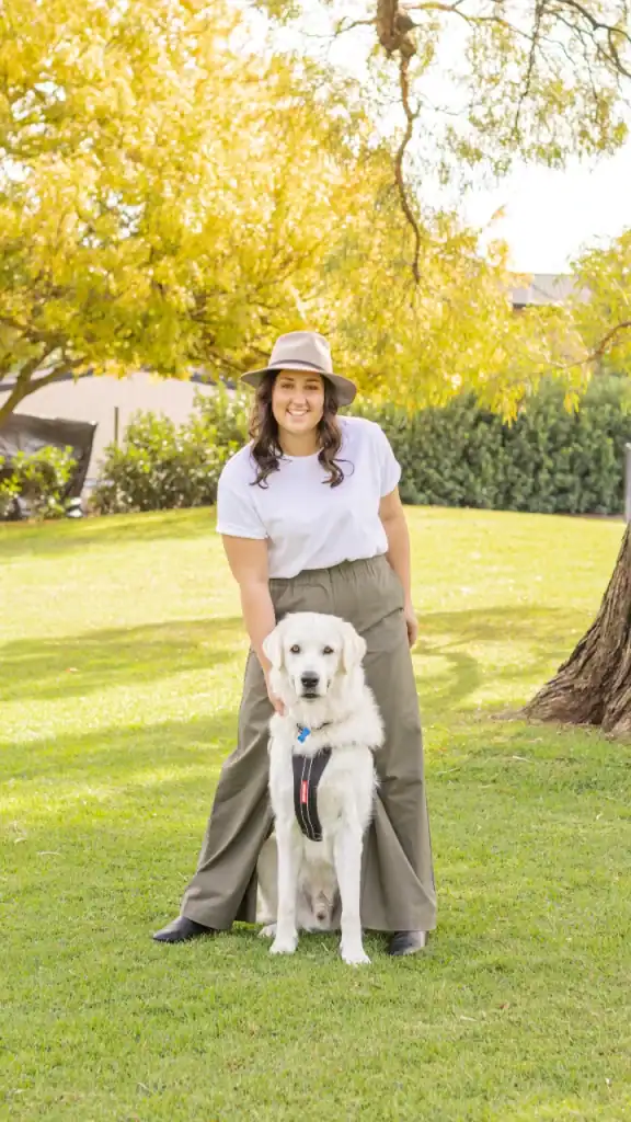 A woman wearing a white shirt, wide-leg olive pants, and a beige hat stands on grass with a golden retriever sitting in front of her. Trees with yellow leaves and sunshine are in the background.
